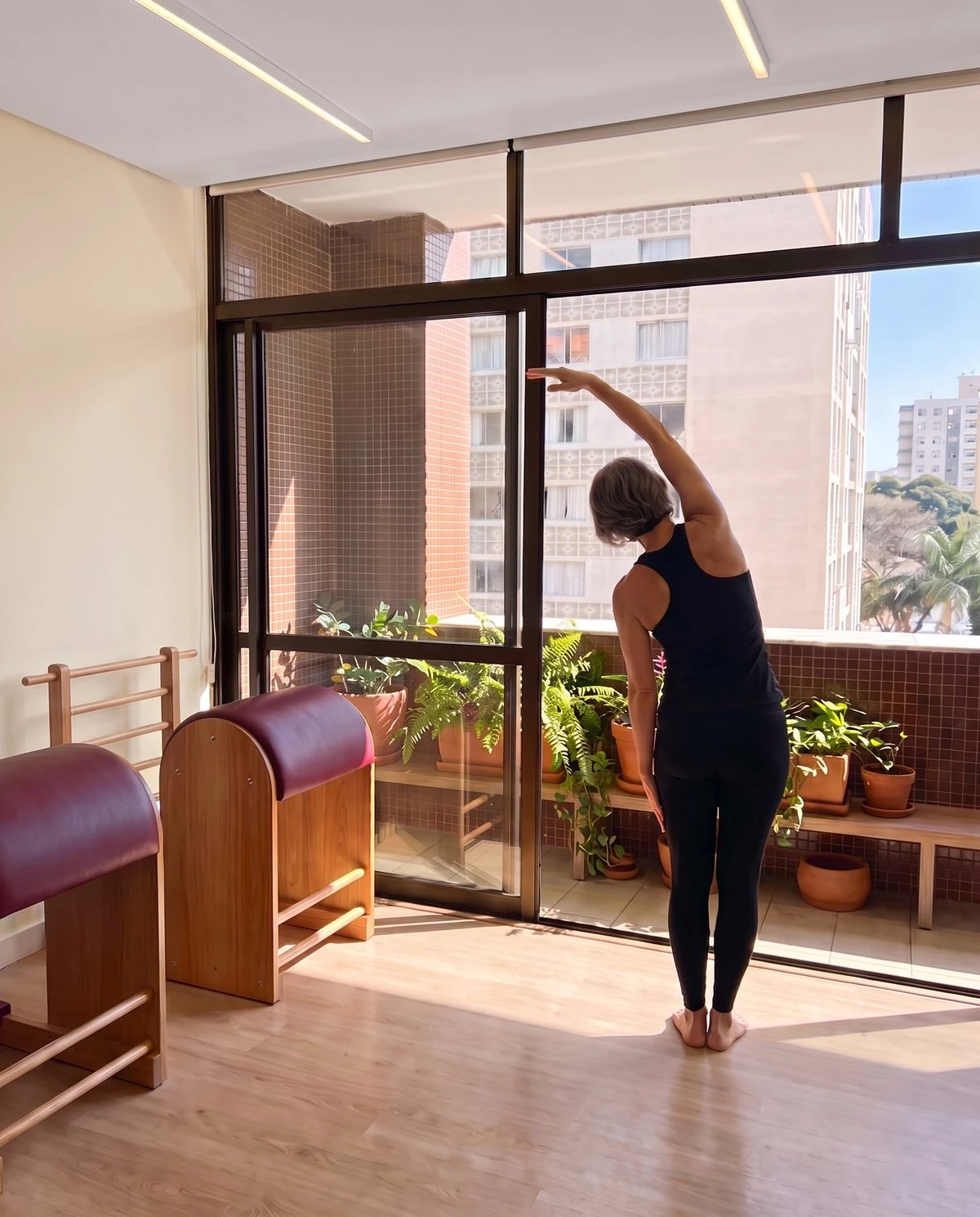 Aluna de costas fazendo um alongamento lateral em pé diante da sacada ensolarada, com plantas e dois Ladder Barrels ao lado